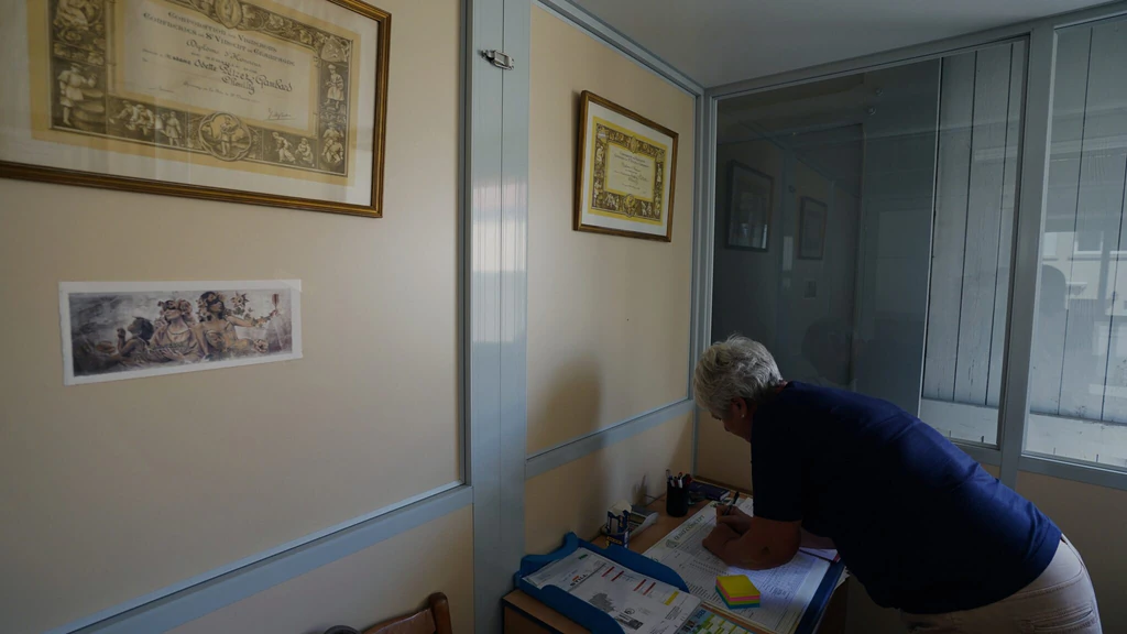 Femme aux cheveux gris penchée sur des documents, dans un bureau orné de diplômes viticoles